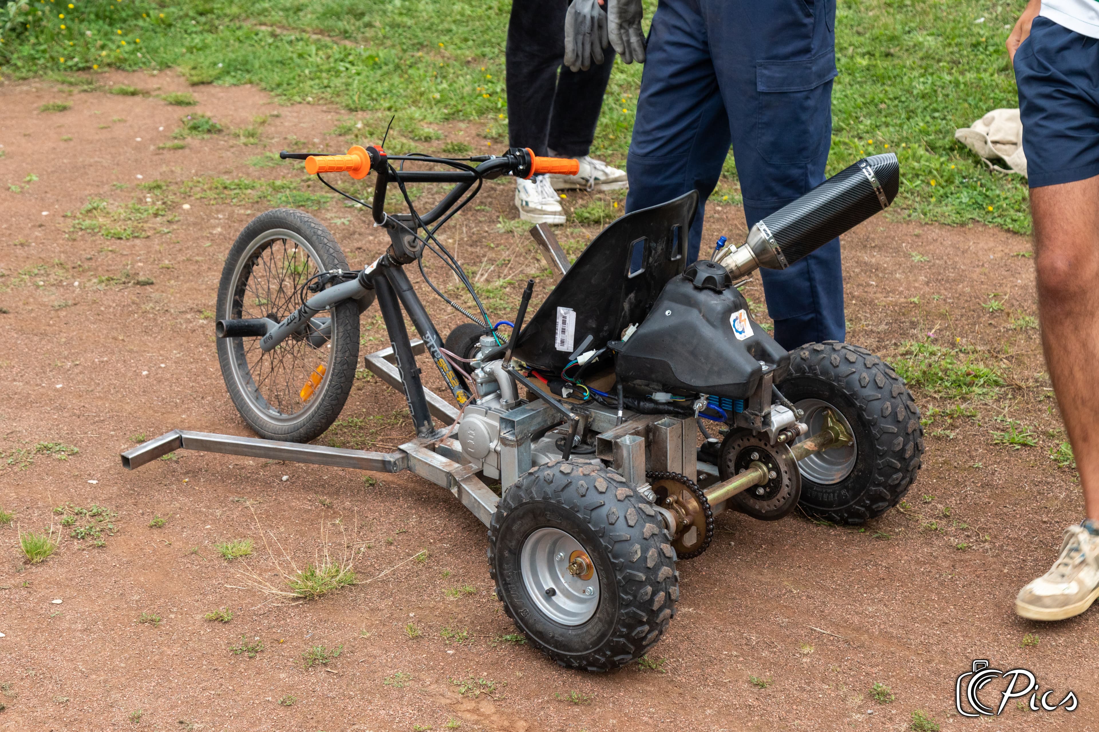 Tricycle motorisé artisanal posé au sol, équipé de roues tout-terrain, d’un cadre métallique et d’un siège baquet, entouré de plusieurs personnes.