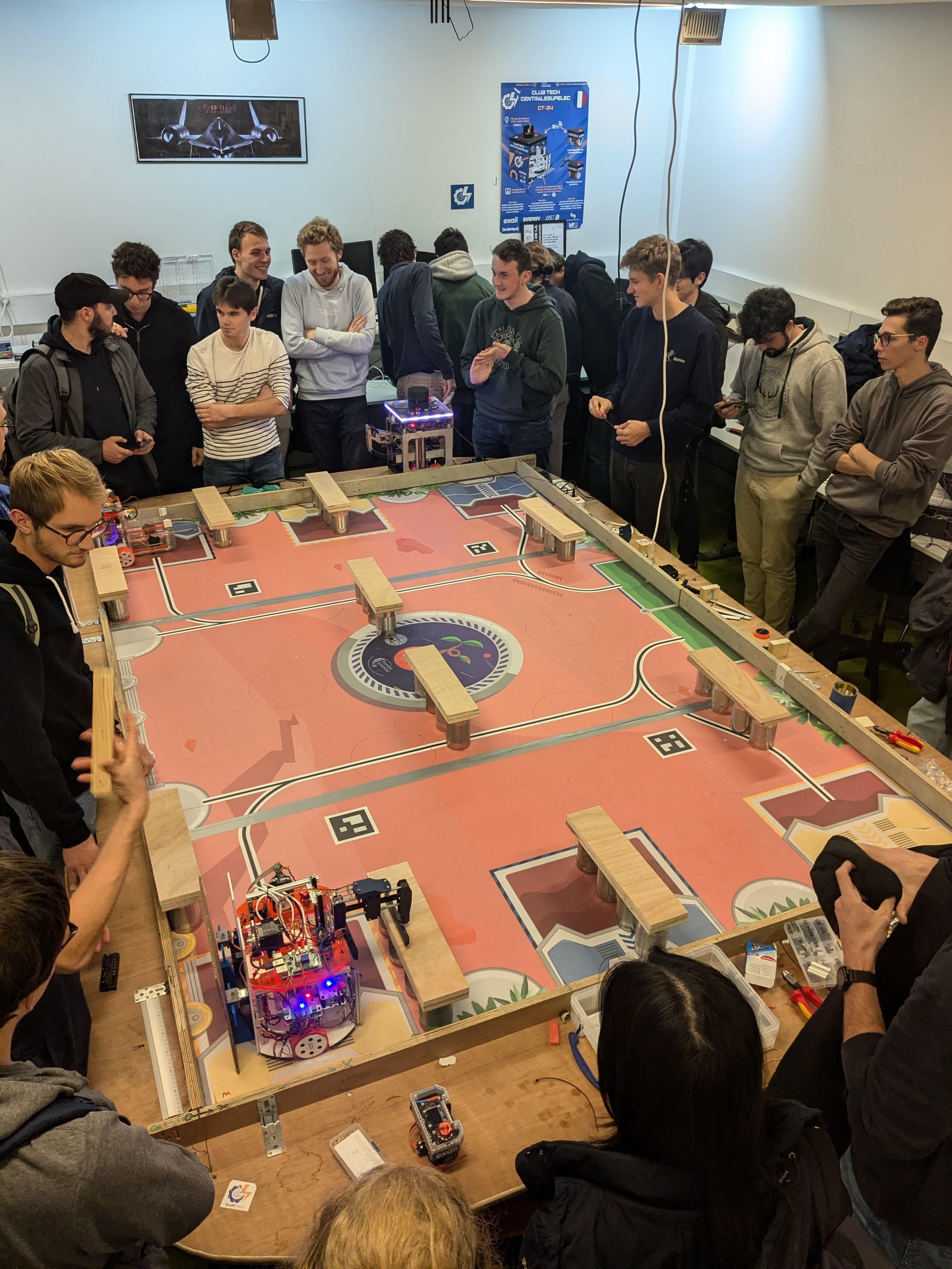 Des étudiants du Club Tech rassemblés autour d’une grande table de jeu colorée, observant deux robots pilotés s’affronter lors du match final du hackathon dans une salle technique.
