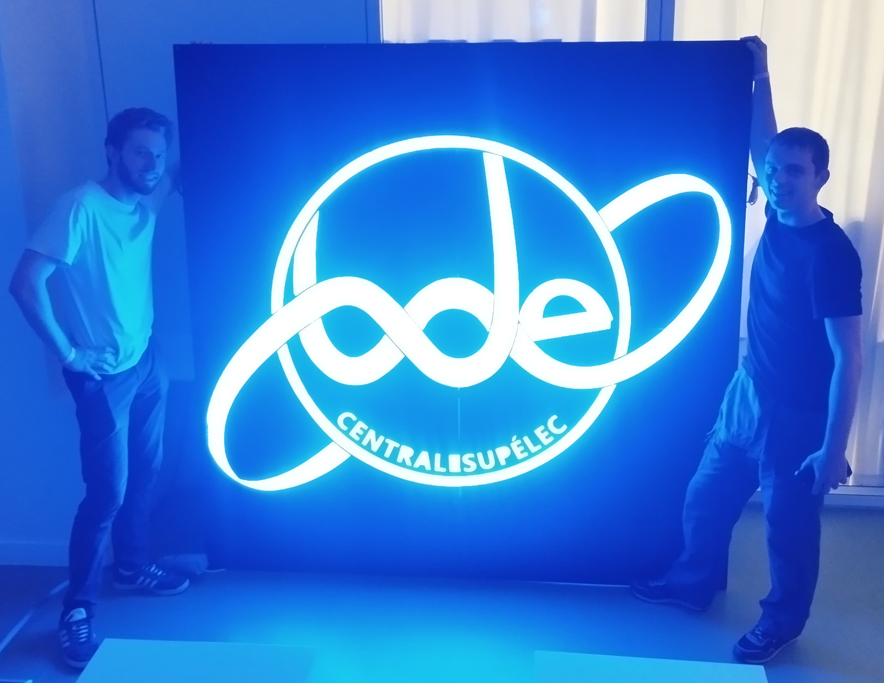 Deux personnes posent de part et d’autre d’un grand panneau lumineux bleu affichant le logo du BDE CentraleSupélec.