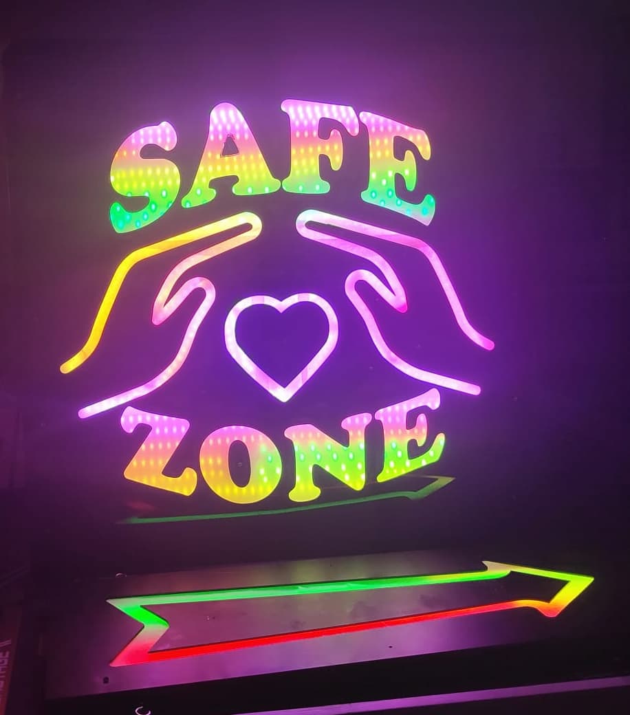 Panneau lumineux aux couleurs arc-en-ciel avec les mots 'SAFE ZONE', un cœur et deux mains stylisées formant une zone de protection.