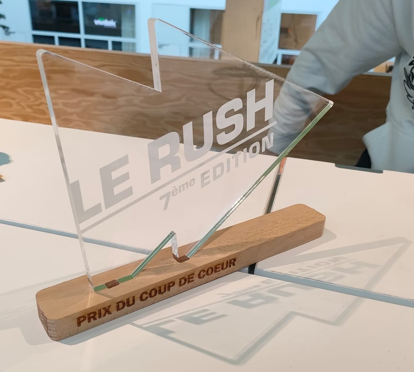 Trophée en verre gravé 'LE RUSH 7e ÉDITION', avec base bois marquée “Prix du coup de cœur”, posé sur une table blanche.