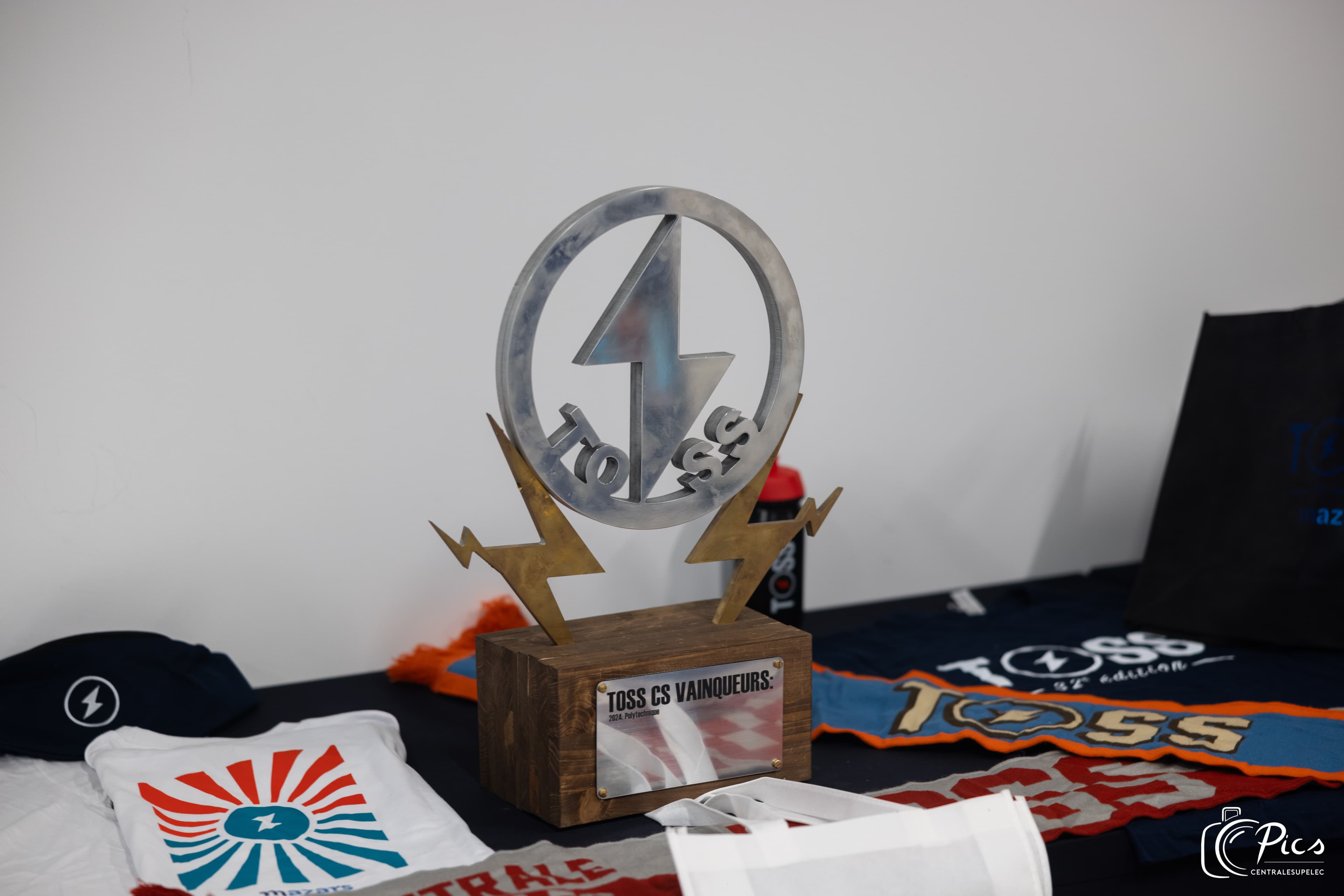 Trophée en métal du TOSS CentraleSupélec avec éclairs et base en bois, posé sur une table avec des vêtements du tournoi.